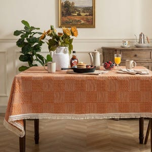 Könnte beinhalten: Eine orangefarbene Tischdecke mit weißem Karomuster und Fransen bedeckt einen Holztisch. Der Tisch ist zum Frühstück gedeckt, mit Pfannkuchen, Obst und Kaffee. Eine Vase mit Sonnenblumen und ein Landschaftsbild befinden sich im Hintergrund.