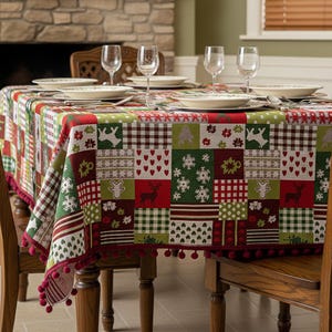 Op de afbeelding: Een feestelijk kerstkleed met een patchwork-ontwerp. Het tafelkleed heeft vierkanten met rendieren, sneeuwvlokken en hartpatronen in rood, groen en wit. De randen zijn afgewerkt met rode pompons. De tafel is gedekt met borden, glazen en bestek.