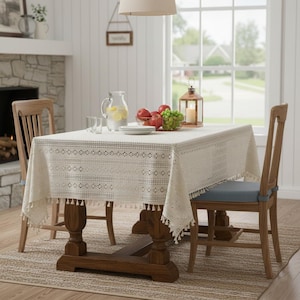 May include: A dining room scene featuring a wooden table set with a cream-colored tablecloth with fringe. The table is set with plates, glasses, and a pitcher of lemonade. Red apples and green grapes are in a wire basket. Two wooden chairs are visible.