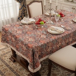 May include: A rectangular tablecloth with a repeating pattern of stylized fan shapes in shades of red, blue, and brown. The tablecloth has a fringed edge and is set on a dark wooden table with place settings, fruit, and silverware.
