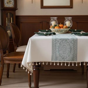 May include: A white tablecloth with a decorative blue-green runner and tassel trim. The table is set with green plates, napkins, and a bowl of pumpkins. Two glass candle holders are on the table.