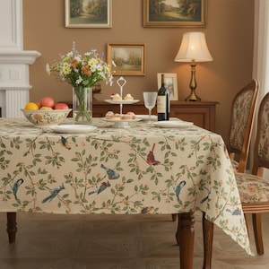 Könnte beinhalten: Beige Tischdecke mit einem sich wiederholenden Muster aus Vögeln und Blättern. Der Tisch ist mit Obst, Blumen, Tellern und Wein gedeckt. Eine Lampe und Stühle sind ebenfalls im Bild zu sehen.