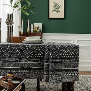 May include: A monochrome geometric patterned tablecloth, featuring triangles and dots, draped over a table. White plates and candles adorn the table, creating a stylish dining setting. The tablecloth's design adds a modern touch to the dining area.