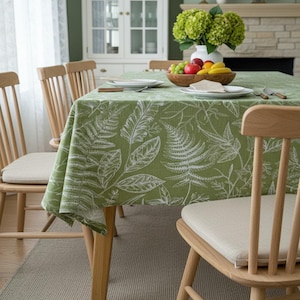 May include: A green tablecloth with a white fern leaf pattern covers a wooden dining table. The table is set with white plates, silverware, and a wooden bowl of fruit. Wooden chairs with beige cushions surround the table. A vase of green flowers sits in the background.