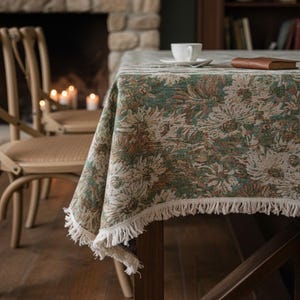 Puede incluir: Un mantel rectangular con un estampado floral en tonos verdes, beige y marrón. El mantel tiene un borde con flecos y está colocado sobre una mesa de madera. Una taza y un platillo blancos están sobre la mesa.