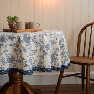 May include: A round tablecloth with a blue floral pattern and a navy blue lace trim. The table is set with mugs, a wooden tray, and a small potted plant. A wooden chair is positioned to the right of the table.
