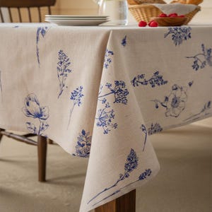 Könnte beinhalten: Eine beige Leinentischdecke mit blauem Blumenmuster. Die Tischdecke liegt auf einem Holztisch, im Hintergrund sind ein Korb mit Himbeeren und ein Stapel Teller zu sehen. Das florale Design umfasst verschiedene botanische Illustrationen.