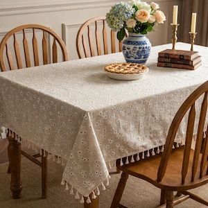 May include: A rectangular beige tablecloth with a delicate floral pattern and tassel trim, covering a wooden table. The table is set with a pie, a blue and white vase with flowers, candles, and books. Wooden chairs surround the table.