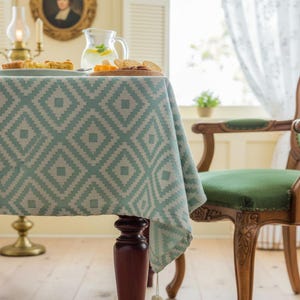 May include: A teal and white geometric patterned tablecloth covers a wooden table. The tablecloth has a diamond design. A wooden chair with a green seat is to the right. A pitcher of lemonade and food are on the table.