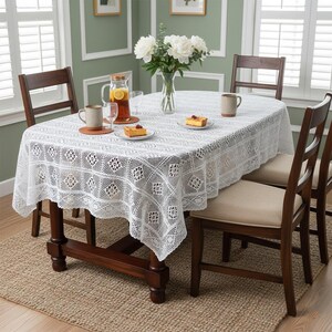 May include: A white lace tablecloth covers an oval wooden table set for a meal. The table is surrounded by wooden chairs with beige cushions. A vase of white flowers and a pitcher of lemonade are on the table.