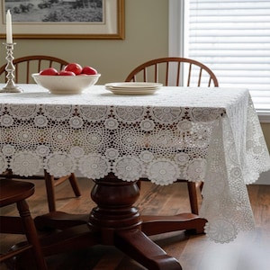 Puede incluir: Un mantel de encaje blanco con un patrón circular cubre una mesa de madera. La mesa está puesta con un cuenco de manzanas rojas y platos. También se ven un candelabro plateado y sillas de madera.