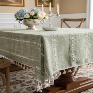 May include: Sage green rectangular tablecloth with white embroidered details and tassel trim. The tablecloth is on a wooden table, with a floral centerpiece and candles in the background. The table is set for a meal.