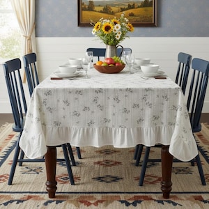 May include: A dining table set for a meal, featuring a white tablecloth with a floral pattern and a ruffled edge. The table is set with white dishes, glasses, and silverware. Blue chairs surround the table, and a vase of sunflowers sits in the center.