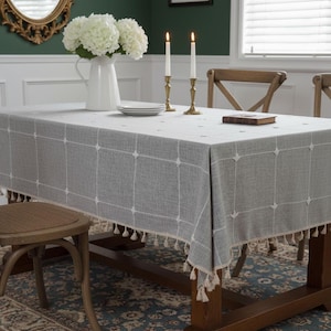 May include: A grey rectangular tablecloth with a white grid pattern and tassel trim. The table is set with white plates, candles, and a vase of white flowers. The table is made of wood.