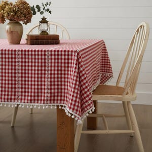May include: A red and white checkered tablecloth with a white pom-pom trim. The tablecloth is draped over a wooden table, with a white chair in the background. A vase of dried flowers and a small lantern sit on the table.