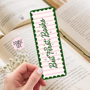 May include: A double-sided bookmark with the text "But First, Books" in a whimsical script. The bookmark has a pink and white striped background with green scalloped edges and star accents. The bookmark is placed in an open book.