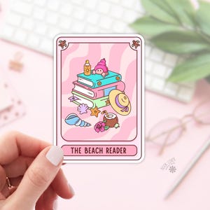 May include: A pink sticker with a cartoon illustration of a person peeking over books, a beach hat, and a coconut drink. The sticker has a pink wavy background and the text "THE BEACH READER".