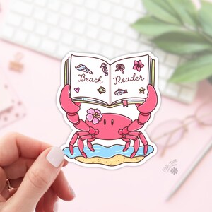 Op de afbeelding: Een roze krabsticker met een boek met de tekst "Beach Reader". De krab heeft een bloem op zijn hoofd en staat op een strand met blauw water. De sticker heeft een witte rand en de tekst "Book Lover" staat onderaan.