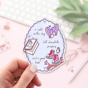 May include: A die-cut sticker with a light purple scalloped border featuring illustrations of purple mittens, a book with a snowman on the cover, pink ice skates, and a penguin. The text reads: a cold winter day, hot chocolate seasons, and a good book.