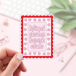 Puede incluir: Una pegatina con un borde festoneado rojo y un interior rosa. La pegatina dice "blind date book club" en una fuente caprichosa, rodeada de pequeños corazones rosas. La pegatina está sujeta por una mano.