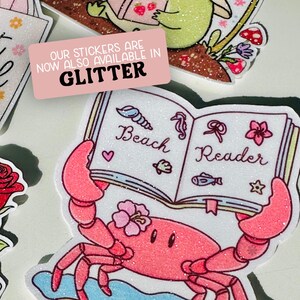 Może przedstawiać: Kolekcja brokatowych naklejek. Jedna naklejka przedstawia r&oacute;żowego kraba trzymającego książkę z napisem "Beach Reader". Naklejki są dostępne w brokatowym wykończeniu, co wskazuje r&oacute;żowy prostokąt z białym tekstem.