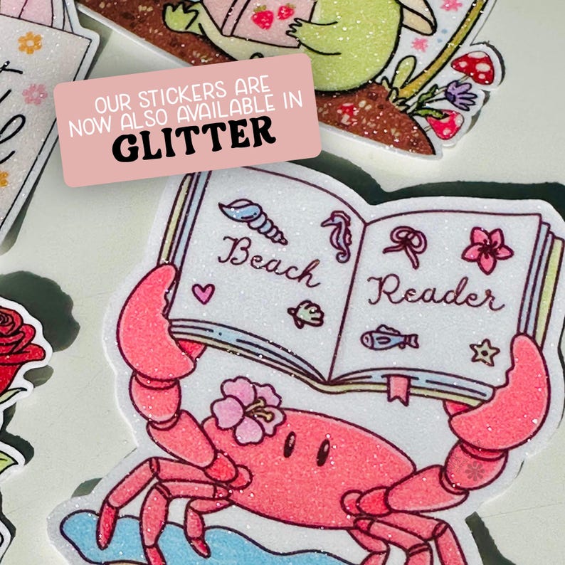 Może przedstawiać: Kolekcja brokatowych naklejek. Jedna naklejka przedstawia r&oacute;żowego kraba trzymającego książkę z napisem "Beach Reader". Naklejki są dostępne w brokatowym wykończeniu, co wskazuje r&oacute;żowy prostokąt z białym tekstem.