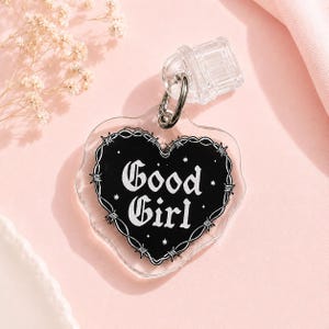 Op de afbeelding: Een hartvormige hanger van helder acryl met een zwart middenstuk en de witte tekst Good Girl in een gotisch lettertype. Het hart is omlijnd met een witte afbeelding van prikkeldraad en kleine steraccenten. Een zilveren metalen ring is bevestigd.