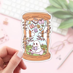 May include: A whimsical sticker featuring an hourglass design with a ghost reading a book inside. The hourglass is adorned with flowers and books, with a bird and a heart. The sticker has a light pink background.