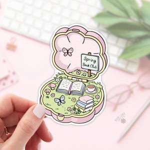 May include: A pink and yellow sticker depicting a book club scene within a clamshell design. The scene includes an open book, a teacup, and a sign reading "Spring Book Club." Butterflies and flowers are also present.