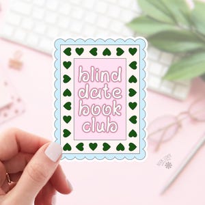 Puede incluir: Una pegatina rectangular con un borde festoneado azul claro y detalles de corazones verdes. La pegatina tiene un centro rosa con las palabras "blind date book club" en una fuente caprichosa. La pegatina está sujeta por una mano.