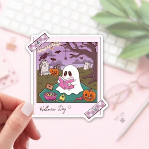 May include: A square sticker designed like a Polaroid photo featuring a white ghost reading a pink book in a graveyard. The scene includes pumpkins, a bowl of candy, and a tombstone under a purple sky. The text Halloween Day is written at the bottom.