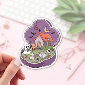 May include: A die-cut sticker featuring a purple, open, clover-shaped compact case containing a miniature Halloween scene. The scene includes a house with an orange roof, a white ghost reading a book, a tombstone marked RIP, a cauldron, pumpkins, a skull, bats, and a crescent moon.