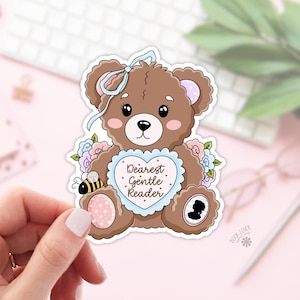 Peut inclure: Autocollant en forme d'ours en peluche marron avec un nœud bleu, des joues roses et un signe en forme de cœur avec l'inscription "Dearest Gentle Reader". L'autocollant comporte également une petite abeille et des fleurs. L'autocollant est tenu par une main.