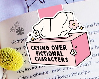 May include: A pink and white bookmark with the text "CRYING OVER FICTIONAL CHARACTERS" on a tissue box design. The bookmark is placed on an open book with the words "BOOK LOVER merch shop" visible.