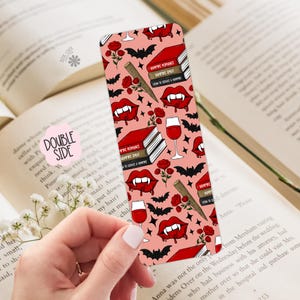 May include: A double-sided bookmark with a pink background featuring a repeating pattern of vampire-themed elements. The design includes red lips with fangs, bats, roses, books, and wine glasses. The text "Vampire Romance" is visible on the books.