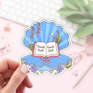 May include: A sticker featuring a blue seashell with an open book that reads "Beach Reads Book Club." The design includes aquatic plants, bubbles, and small flowers. The sticker is held by a person's hand.