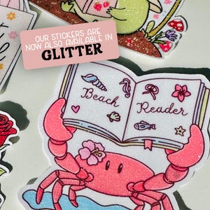 May include: A collection of glitter stickers featuring a crab holding a book that says "Beach Reader". The crab is pink with a flower on its head. The stickers are available in glitter, as indicated by the pink rectangle with white text.