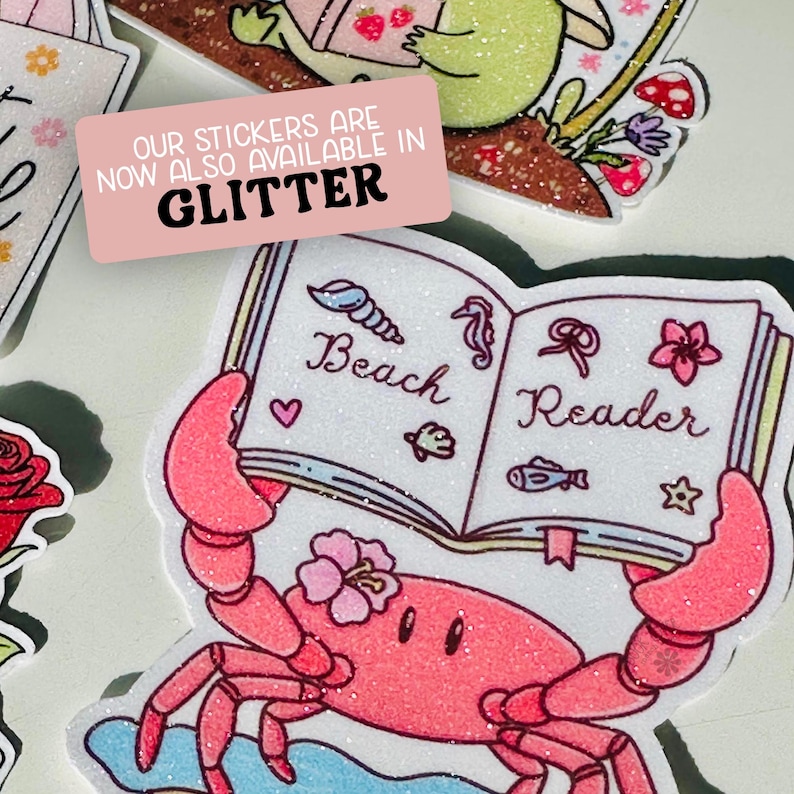 May include: A collection of glitter stickers featuring a crab holding a book that says "Beach Reader". The crab is pink with a flower on its head. The stickers are available in glitter, as indicated by the pink rectangle with white text.