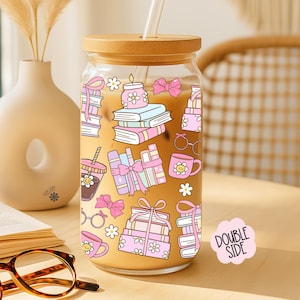 May include: A clear glass tumbler with a bamboo lid and straw, decorated with pink and light blue book-themed illustrations. The tumbler has a double-sided design and is filled with a light-colored beverage. A vase and eyeglasses are in the background.