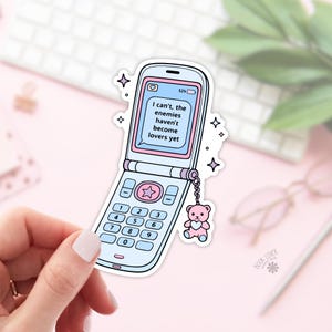 Puede incluir: Una pegatina con un diseño de teléfono plegable azul claro con una pantalla y detalles en rosa. La pantalla muestra el texto "I can't, the enemies haven't become lovers yet." Un pequeño dije de oso de peluche rosa cuelga del teléfono.