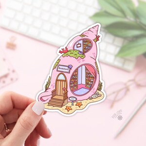 May include: A pink seashell-shaped sticker depicting a bookstore. The design includes a door, windows, and shelves filled with books. The word "Bookstore" is printed on the side. The sticker is set on a sandy beach with starfish.