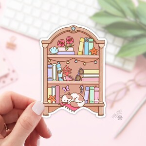 May include: A sticker featuring a bookshelf design with books, flowers, and sea-themed decorations. A sleeping cat rests on a pink pillow at the bottom. The sticker is held by a hand against a pink background.
