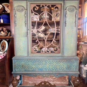 Op de afbeelding: Een vintage, teal-kleurige kast met een glazen deur en sierlijke metalen accenten. De kast heeft een verweerde afwerking en een decoratief stencilafdruk op de lade. De kast staat op vier poten met een decoratieve basis.