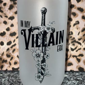 Peut inclure: Gobelet en verre givré en forme de canette avec un couvercle en bois. Le gobelet présente un motif d'épée noire entrelacée de fleurs et les mots "IN MY VILLAIN ERA" en lettres noires.