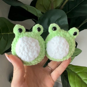 May include: Two green crocheted frog plushies with white bellies and black eyes. The plushies are being held in a person's hand.