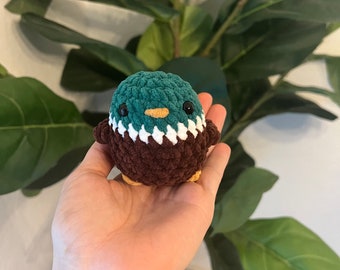 May include: A small crocheted stuffed animal shaped like a bird. The bird is brown, green, and white with black eyes and an orange beak.
