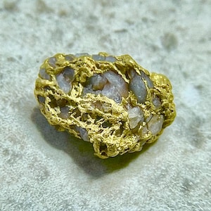 Puede incluir: Una pepita de oro natural con una superficie metálica amarilla texturizada e inclusiones minerales de color gris claro translúcido. El espécimen presenta una forma orgánica irregular con una estructura porosa de oro en forma de red.