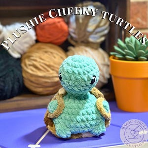 May include: A handmade plush turtle in shades of teal and brown, with black eyes. The turtle is crocheted and sits on a purple surface. The image includes the text "PLUSHIE CHEEKY TURTLES" and a logo that says "MAGIC CIRCLE CREATIONS".