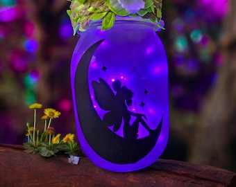 May include: A glass jar with a purple glow and a silhouette of a fairy sitting on a crescent moon. The jar is decorated with a green and brown twine bow and white flowers.