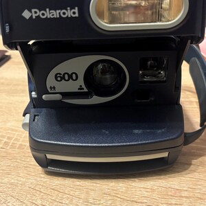 May include: A navy blue Polaroid 600 instant camera with a built-in flash. The camera has a black strap and a white Polaroid logo. The lens is centered below the flash, and the number 600 is printed on the front.